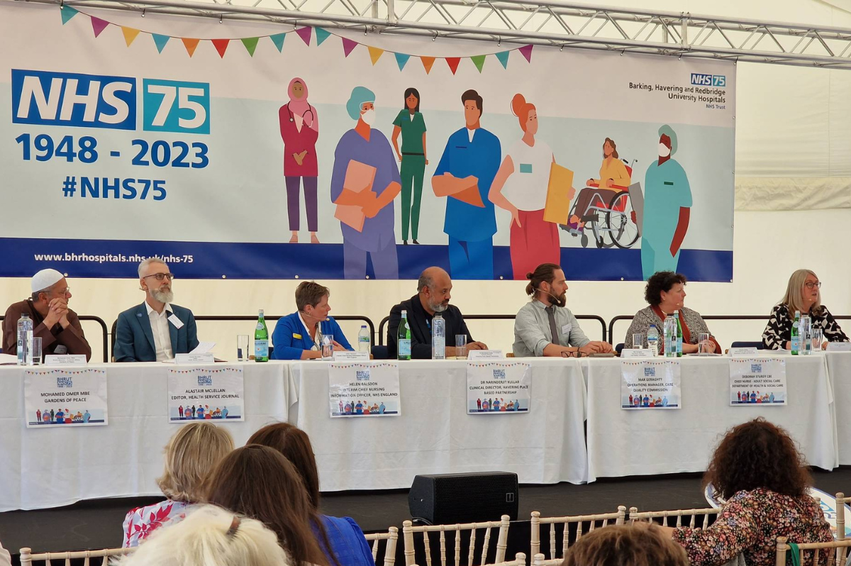 A photo of the panel sat behind a long white table. Behind the panel is a large banner with our NHS 75 design on it